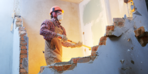 Photo of a contractor in a mask and hard hat sledgehammering through a brick and plaster wall