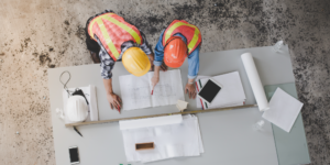 Top-down photo of two workers in hard hats reviewing construction project monitoring documents at a table at a construction site